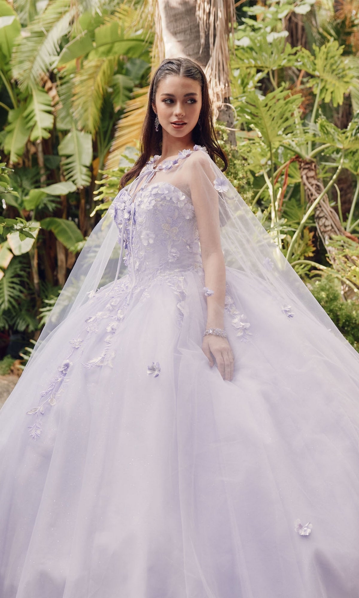 A woman wearing the Juliet 1435 Strapless Ball Gown Quinceañera Dress with floral details and a sheer cape stands outdoors among lush green tropical plants.